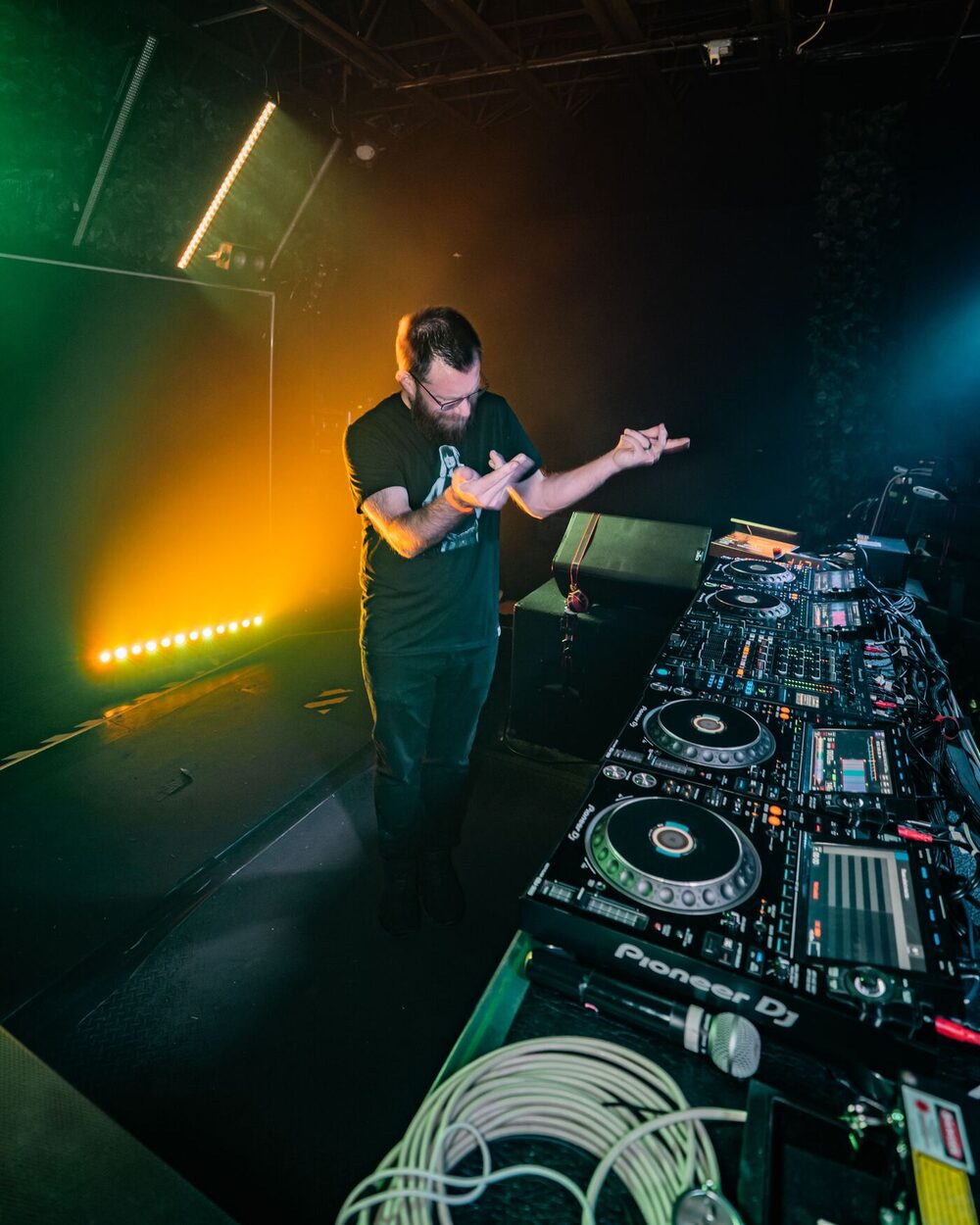 Sam behind a Pioneer DJ setup on a smoke-filled stage lit green and amber.